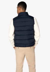 Hombre de espaldas con un chaleco acolchado azul marino sobre una camisa blanca de manga larga y pantalones beige, con las manos en los bolsillos.