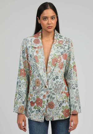 Femme aux longs cheveux noirs portant un blazer à bouton unique orné d'une broderie florale multicolore sur un fond clair.