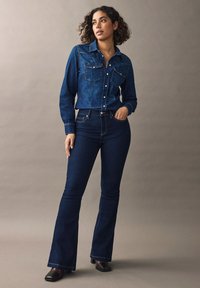 Cămașă din denim cu buzunare frontale și închidere cu nasturi, asociată cu blugi de tip flared de culoare albastru închis cu talie înaltă. Ambele articole prezintă cusături contrastante.