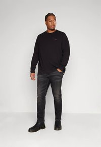 Juoda ilgomis rankovėmis marškinėliai su mažu logotipu, derinami su tamsiais džinsais ir juodais aulinukais. Modelis stovi su rankomis kišenėse.