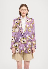 Lilla blomsterblazer med grønne og pink accenter, der har to frontlommer og et skræddersyet design. Båret over en hvid skjorte og beige shorts.