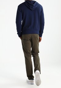 Marinblå hoodie med dragsko och känguruficka, kombinerad med olivgröna byxor och vita sneakers med texturerade sulor.