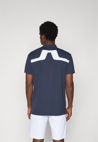 Polo de marina cu manșete scurte, având un design geometric mare alb pe partea din spate. Textură netedă, fabricat dintr-un material ușor.