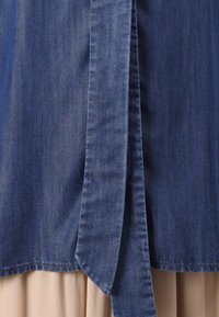 Dunkelblaues Denim-Oberteil mit einem langen Bindeband in der Taille; hat eine glatte Textur und einen geraden Saum, über einem beigen Stoff getragen.