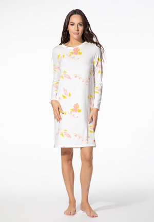 Frau steht barfuß und trägt ein weißes langärmeliges Kleid mit pinken und gelben Blumenmustern auf einem schlichten weißen Hintergrund.
