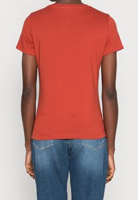 Persona vistiendo una camiseta roja de manga corta lisa y jeans de mezclilla azules, mostrada desde la parte de atrás contra un fondo liso.
