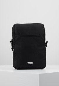 Černá obdélníková taška vyrobená z texturované látky. Má zipové zavírání, nastavitelné ramenní popruhy a bílé logo Levi's na přední straně.