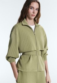 Femme aux longs cheveux portant une veste zippée vert clair à fines rayures avec ceinture et pantalon assorti, debout avec une main dans la poche.