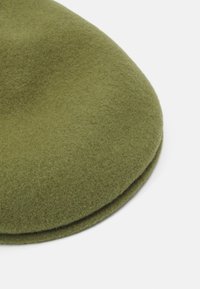 Bonnet en laine vert olive avec une texture lisse, une forme arrondie et un revers replié. Le tissu semble dense et chaud, adapté aux temps froids.
