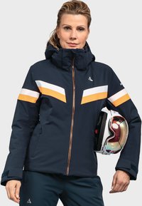 Marineblaue Skijacke mit orangefarbenen und weißen horizontalen Streifen, mit Reißverschluss, verstellbarer Kapuze und getragen mit einem weißen Helm. Robustes, isolierendes Material.