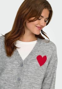 Cardigan grigio realizzato in morbido tessuto knit, con un accento a forma di cuore rosso lavorato a maglia sul lato sinistro, scollatura a V e chiusura con bottoni.
