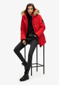 Parka roja con capucha de pelo sintético, cierre de cremallera y botones a presión, combinada con suéter negro, jeans negros y botas de combate negras con cordones.