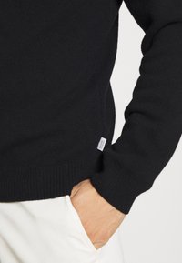 Suéter negro de punto con un cuello redondo acanalado y puños. Presenta una pequeña etiqueta en la parte inferior izquierda. Textura suave, diseño ajustado.