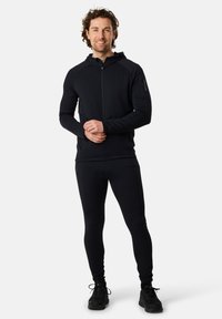 Giacca atletica nera con zip e cappuccio, realizzata in tessuto strutturato, abbinata a pantaloni aderenti dello stesso colore e scarpe da ginnastica nere.