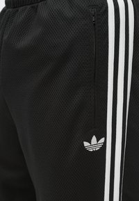 Μαύρο αθλητικό παντελόνι από πλέγμα με λευκές κάθετες ρίγες και μικρό λευκό λογότυπο στην κάτω αριστερή πλευρά. Διαθέτει τσέπη με φερμουάρ.