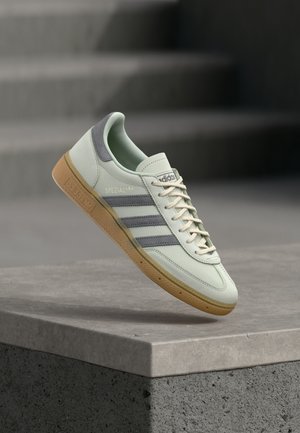 Baskets Adidas vert clair avec des rayures grises et une semelle en gomme flottant au-dessus d'un rebord en béton et d'escaliers en arrière-plan.