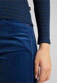 Haut à manches longues bleu côtelé avec des rayures vertes, associé à une jupe en velours côtelé bleu avec une poche et un détail de bouton en métal.