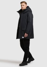 Czarna parka z kapturem, z zapięciem na zamek, dużymi kieszeniami z przodu i naszywką z logo. W połączeniu z czarnymi spodniami cargo i butami.