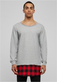 Sweatshirt gris à manches longues en mélange de coton, avec un col large et une couche inférieure à carreaux noir et rouge portée sur des leggings noirs.