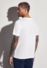 Muž s tetováním na celých rukou, oblečený v jednoduchém bílém tričku a tmavých kalhotách, dívající se do strany před bílou zdí se stínem.