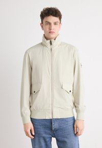 Lys beige zip-up jakke med høy krage, to frontlommer med klaff og knappefeste, samt elastiske mansjetter, kombinert med blå jeans.