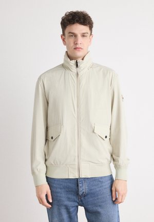 Light beige zip-up jacket, featuring a high collar, two front flap pockets with button closures, and elastic cuffs, paired with blue jeans.