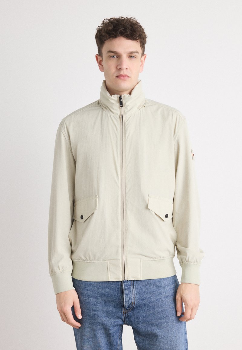 Lys beige zip-up jakke med høy krage, to frontlommer med klaff og knappefeste, samt elastiske mansjetter, kombinert med blå jeans.