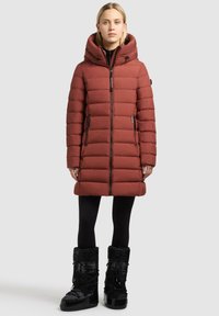 Cappotto puffy rosso con colletto alto e cappuccio regolabile. Presenta tasche con zip e una cerniera frontale; abbinato a stivali invernali neri isolati.