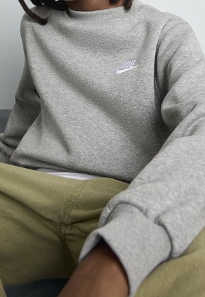 Person iført en grå Nike sweatshirt og olivengrønne bukser, siddende mod en ensfarvet baggrund.