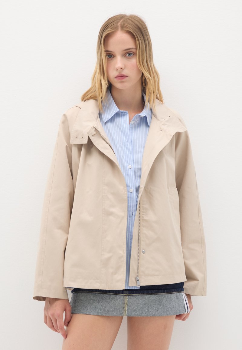 Helles beiges Jacket mit strukturiertem Design, ausgestattet mit einem Reißverschluss, Seitentaschen und einer abnehmbaren Kapuze. Getragen über einem blau gestreiften Hemd.