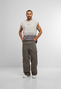 Ärmelloses Tanktop in einem Farbverlauf von Creme zu Grau mit einem Logo auf der Brust. Cargo-Hosen in Dunkelgrau, die mehrere Taschen haben. Weiße Sneaker.