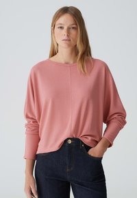 Locker sitzendes, langärmliges Oberteil aus weichem pinken Stoff mit Rundhalsausschnitt und einer zentralen Naht. Kombiniert mit dunklen Jeans.