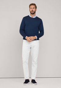 Suéter azul marino sobre una camisa de cuello azul claro, combinado con jeans blancos y zapatillas deportivas negras. Diseño simple, textura suave y ajuste casual.