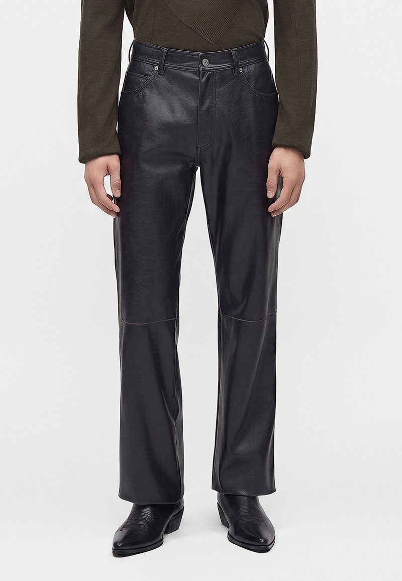 Man wearing black straight-leg leather pants, black cowboy boots, and an olive green long-sleeve top standing against a plain background.