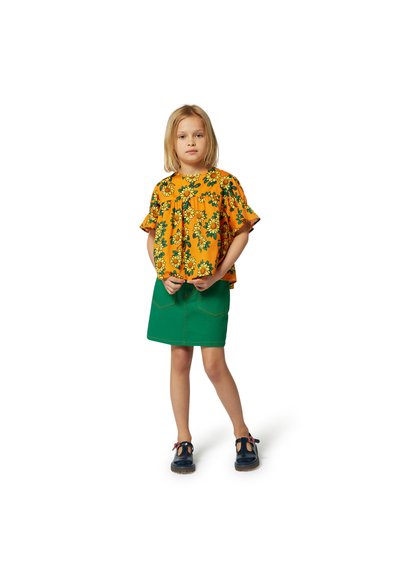 Haut floral jaune avec des manches à volants, associé à une jupe verte. Chaussures noires avec des accents rouges. Tenue simple et décontractée pour enfants.