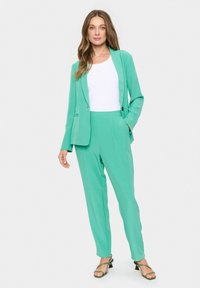 Mint zelena obleka, ki jo sestavljata krojena blazer in ožji hlače, v kombinaciji z belo majico, poudarjena z zelenimi petami.