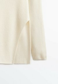 Manche et côté d'un pull en tricot côtelé couleur crème, montrant une texture douce et un léger détail de tissu superposé.