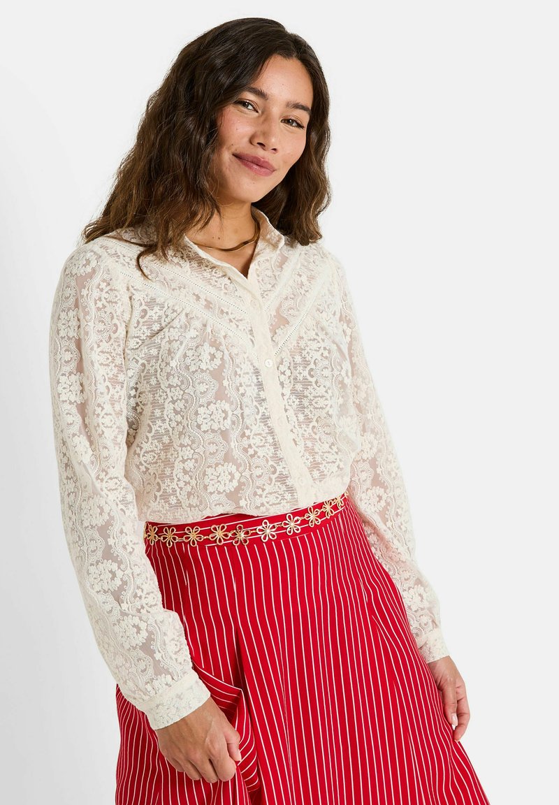 Femme aux cheveux bruns ondulés portant un chemisier en dentelle florale blanche à boutons et une jupe plissée rouge à rayures verticales blanches avec une ceinture chaîne dorée.