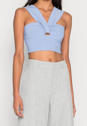 Kobieta ubrana w jasnoniebieski skręcany krótki top i jasnoszare, szerokie spodnie z wysokim stanem, stojąca na jednolitym tle.