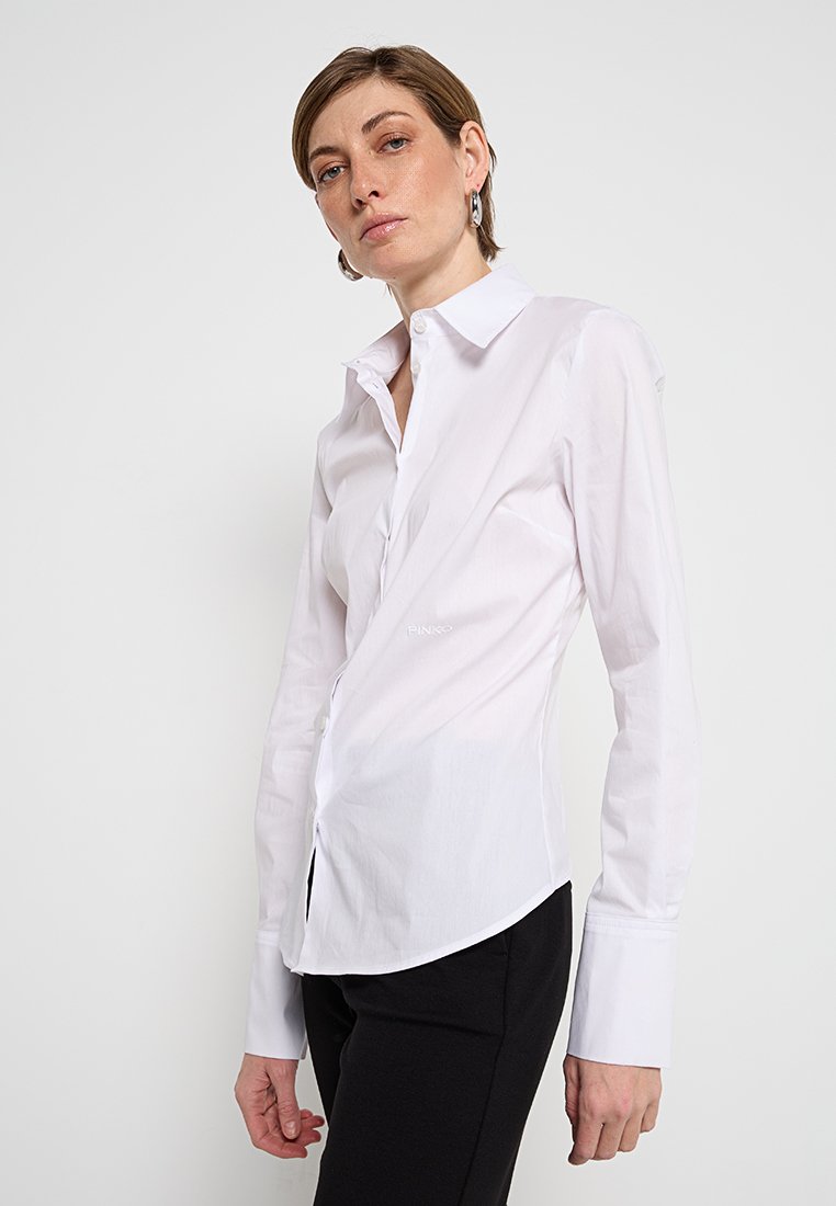 Weißes Baumwollhemd mit einem figurbetonten Design, das einen klassischen Kragen, lange Ärmel und einen abgerundeten Saum aufweist, ergänzt durch Knopfdetails.