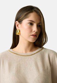 Orecchini dorati e scultorei indossati da una modella, abbinati a un maglione beige con dettaglio al collo simile a una catena. Primo piano, con focus sugli accessori.