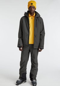 Chaqueta impermeable gris oscuro con capucha, combinada con pantalones gris oscuro. Lleva puesta una camisa amarilla brillante y está acentuada con un gorro amarillo.