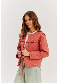 Pikowana, koralowo-różowa kurtka z falbanami, okrągłym dekoltem i długimi rękawami. Posiada sznurki do regulacji i naszywkę z logo na dolnym brzegu.