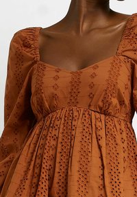 Rostfärgad klänning i strukturerad tyg med broderade detaljer och utskuret mönster, med puffärmar och fyrkantig halsringning.