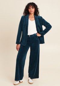 Costume en velours côtelé teal avec un pantalon à jambes larges et un blazer à simple boutonnage, associé à une chemise blanche à col en V et des baskets blanches. Tissu texturé.