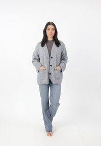 Cardigan grigio chiaro con maglia testurizzata, caratterizzato da un collo a V, chiusura con bottoni, due tasche frontali e spalle scese, abbinato a jeans blu.