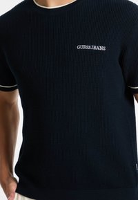 Námořnické pletené tričko s krátkými rukávy, bílé lemování na límci a rukávech, s vyšitým logem "GUESS JEANS" na přední části.