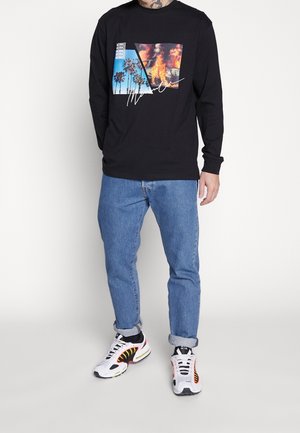 Muž oblečený v černém tričku s dlouhým rukávem s potiskem palem a ohně, světle modré vyhrnuté džíny a bílé tenisky s černými a oranžovými akcenty.