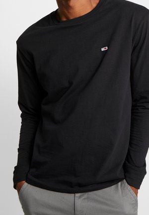 Man draagt een zwarte lange mouwen shirt met een klein rechthoekig logo op de borst en lichtgrijze broek met handen in de zakken.