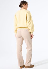 Mujer de pie de espaldas, vistiendo un sudadera amarilla, pantalones de pana beige, zapatos mocasines marrones, cabello recogido en un moño bajo.